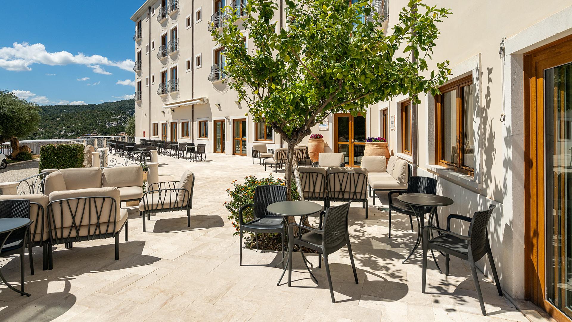 Terrazza assolata con sedie eleganti, alberi e vista collinare.