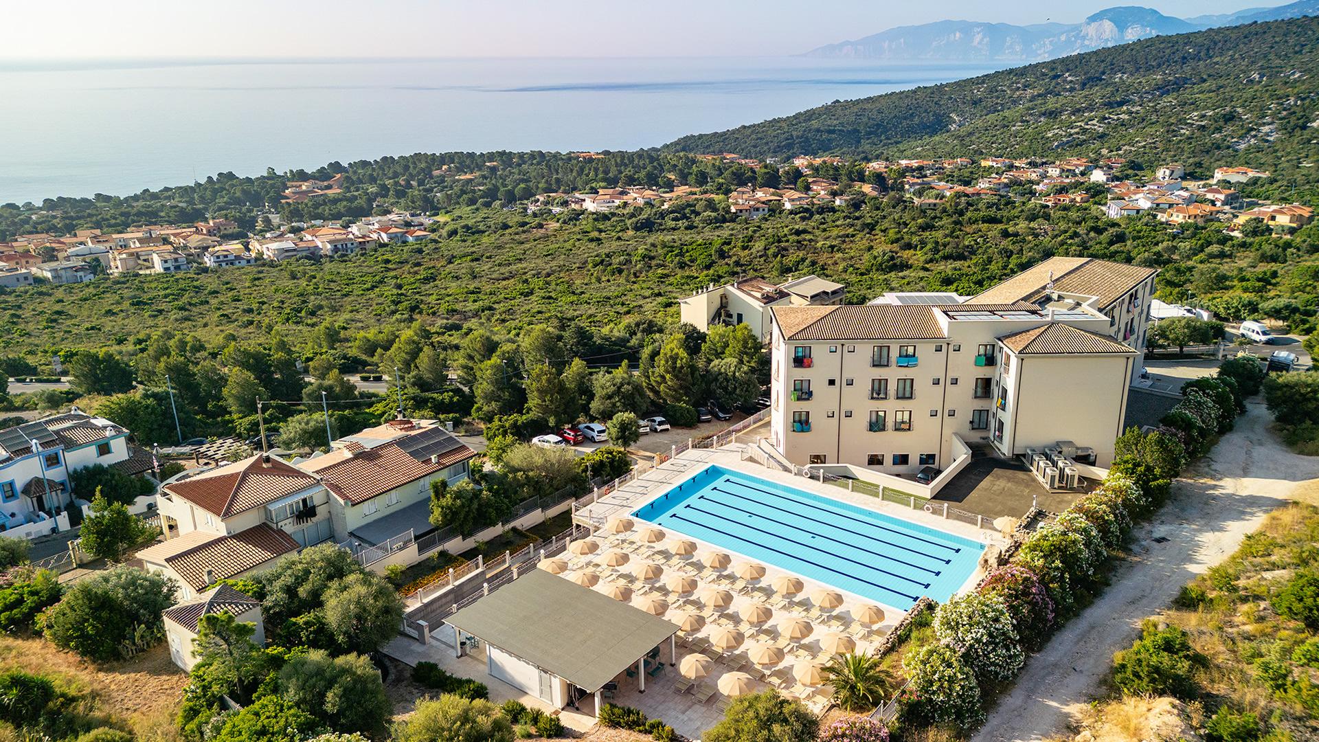 Vista panoramica su hotel con piscina, verde rigoglioso e mare all'orizzonte.